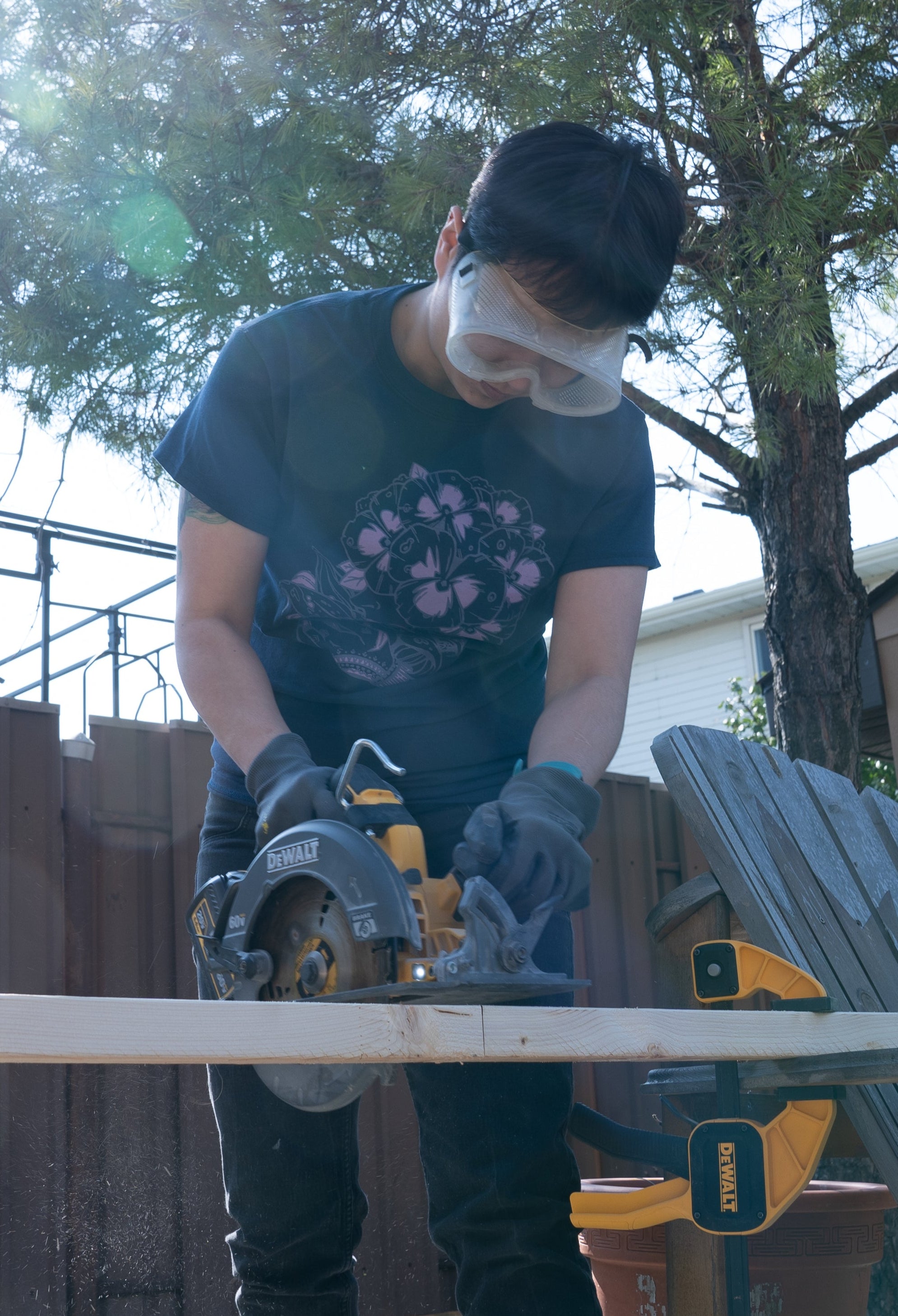 Person using a circular saw on a wooden board outdoors with trees and a fence in the background.