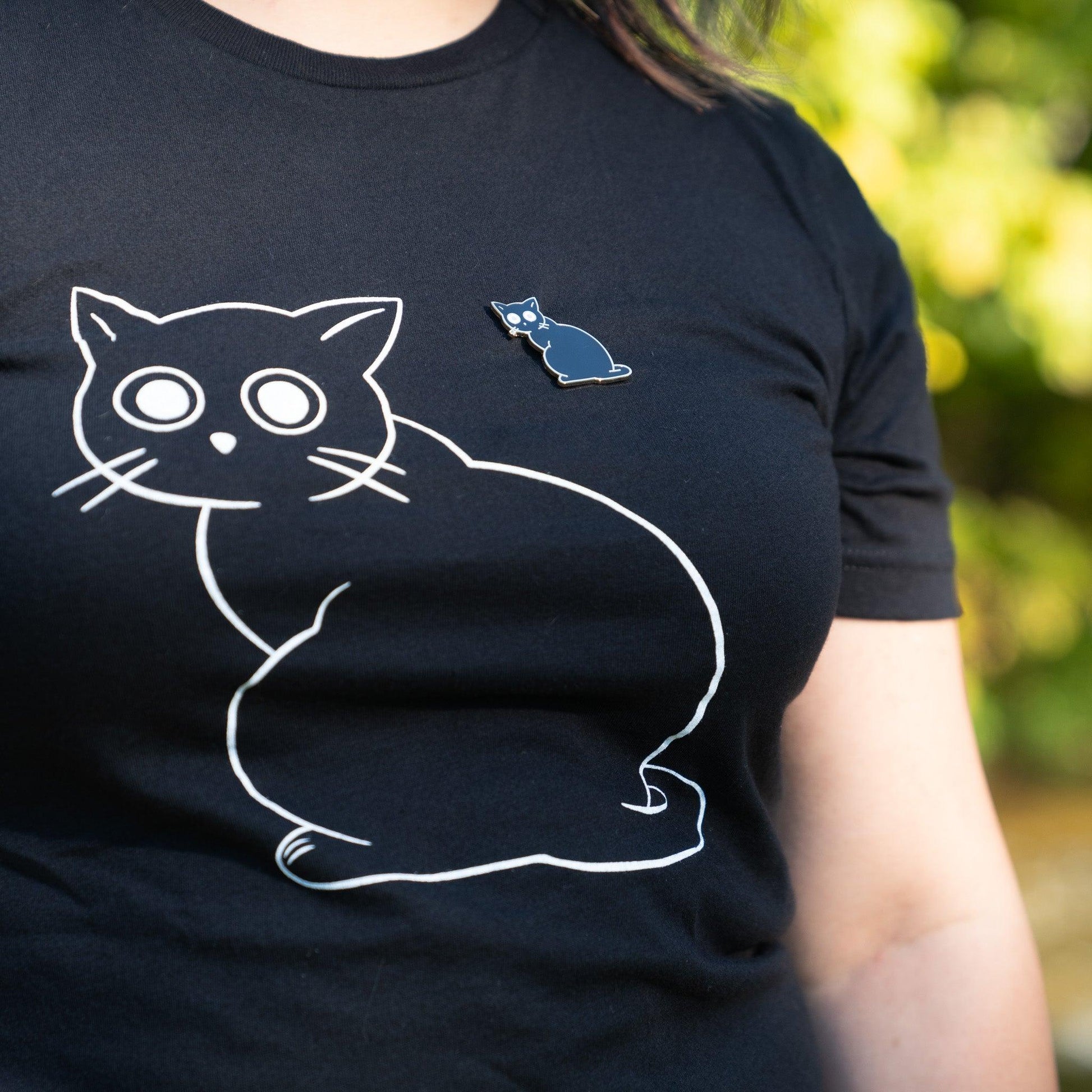 black 100% cotton t-shirt with white linework of a cat with big eyes staring at you. There is a pin of a black cat same image on the shirt as well